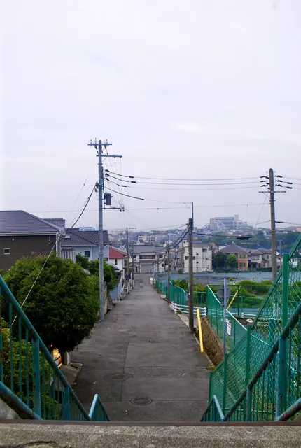 緑のフェンスに沿って、遠くの家並みへ下っていく坂道