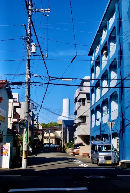 電線が幾重にも交差する青空の下、青い壁の集合住宅が建つ通り