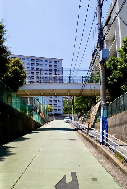 緑色に塗られた坂道の先に、歩道橋と団地が見える風景