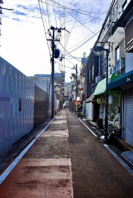 白い工事の囲いと小さな店のあいだを、まっすぐ続く赤い舗装の道