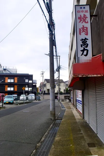 赤い日よけと「焼肉 萬力」の縦看板が立つ、大通り沿いの歩道