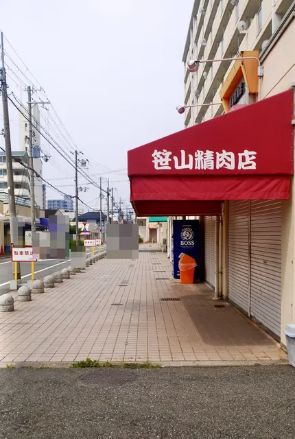 「笹山精肉店」の赤い日よけと、車止めの並ぶ広い歩道