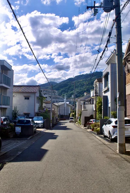 雲の広がる空の下、住宅街の道の先に緑の山が近くに見える風景