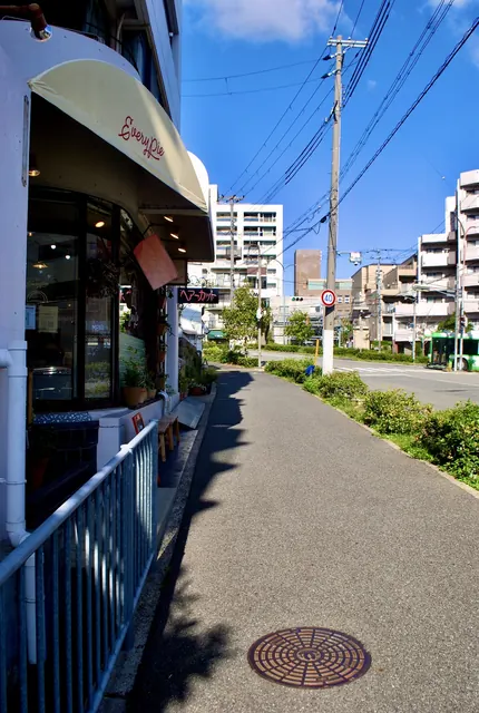 白い日よけのパン屋の店先と、植え込みの続く明るい歩道