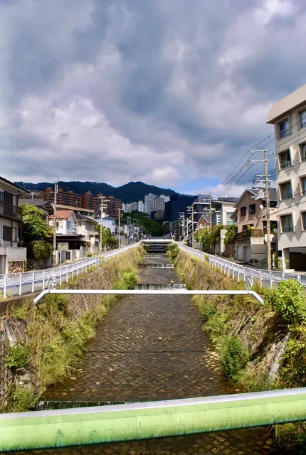 雲の広がる空の下、石積みの川の先に山並みが見える風景