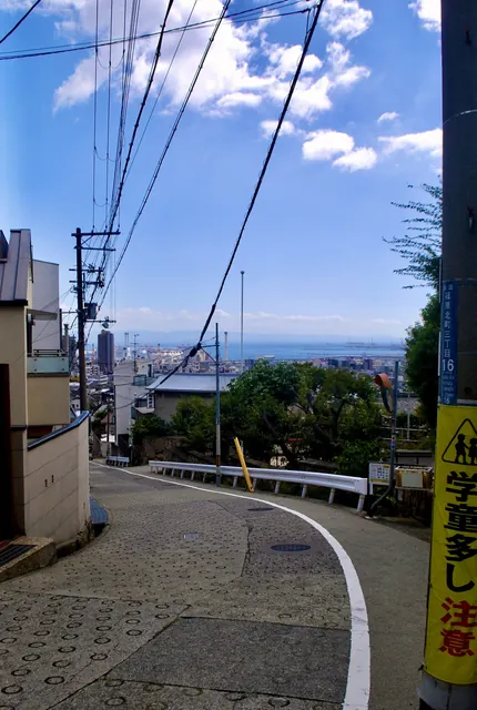 電線の張られた空の下、カーブした坂の先に町と海が広がる風景