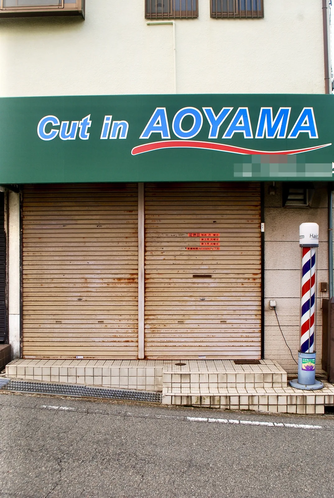 緑の看板とサインポールのある、シャッターの閉まった理容店