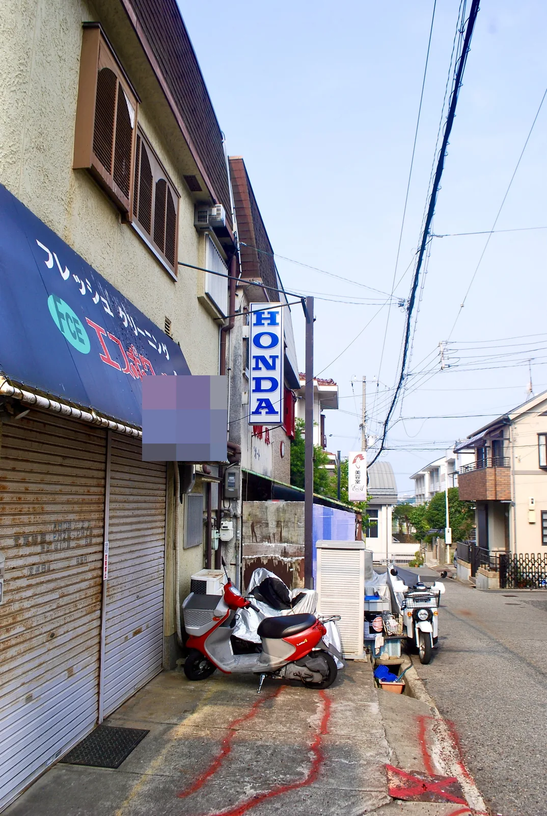 青い日よけのクリーニング店とバイクの並ぶ、奥へ続く細い道