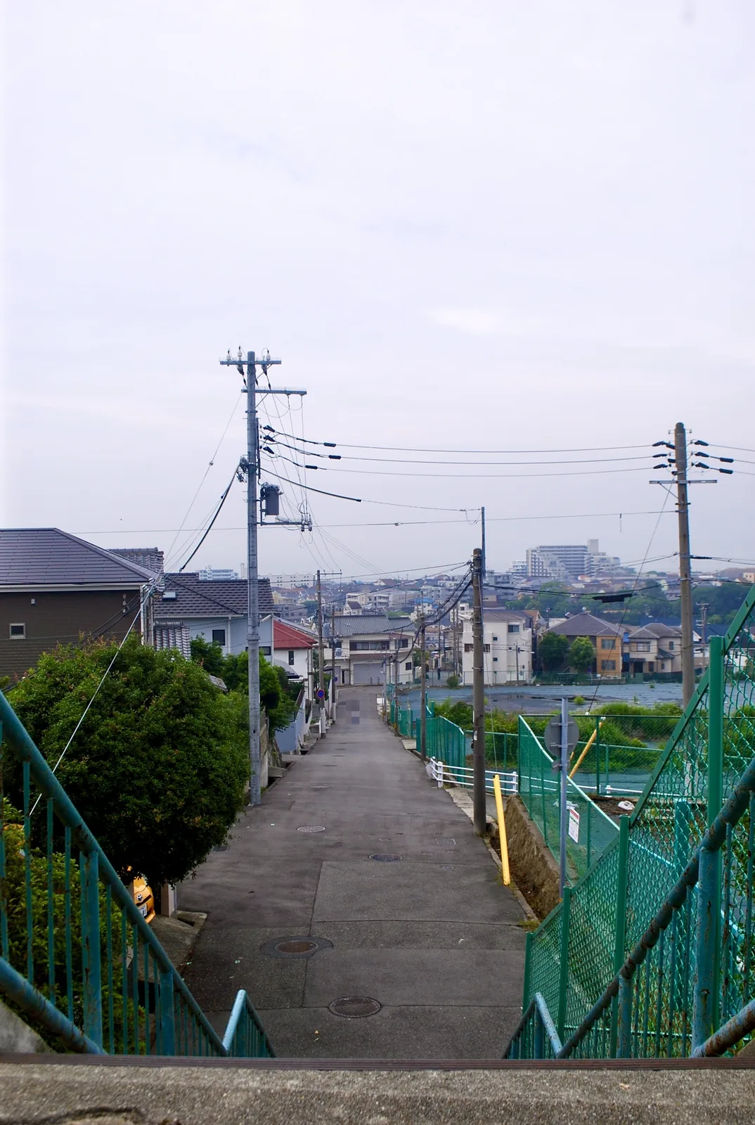 緑のフェンスに沿って、遠くの家並みへ下っていく坂道