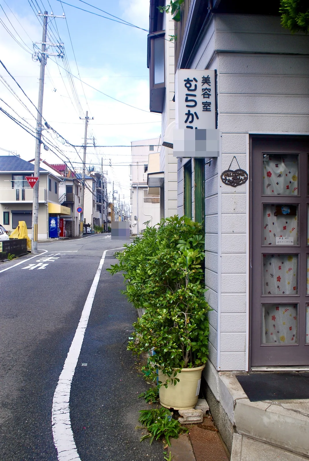 植木と花柄のカーテンの扉、「美容室 むらかみ」の看板がある曲がり角