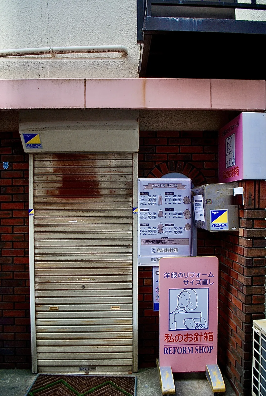 レンガの壁にシャッターと立て看板のある、小さな洋服直しの店