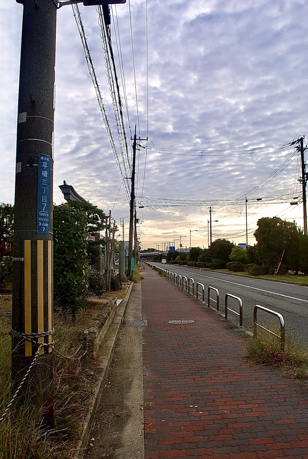 夕方の雲が広がる空の下、柵の並ぶ歩道とまっすぐな大通り