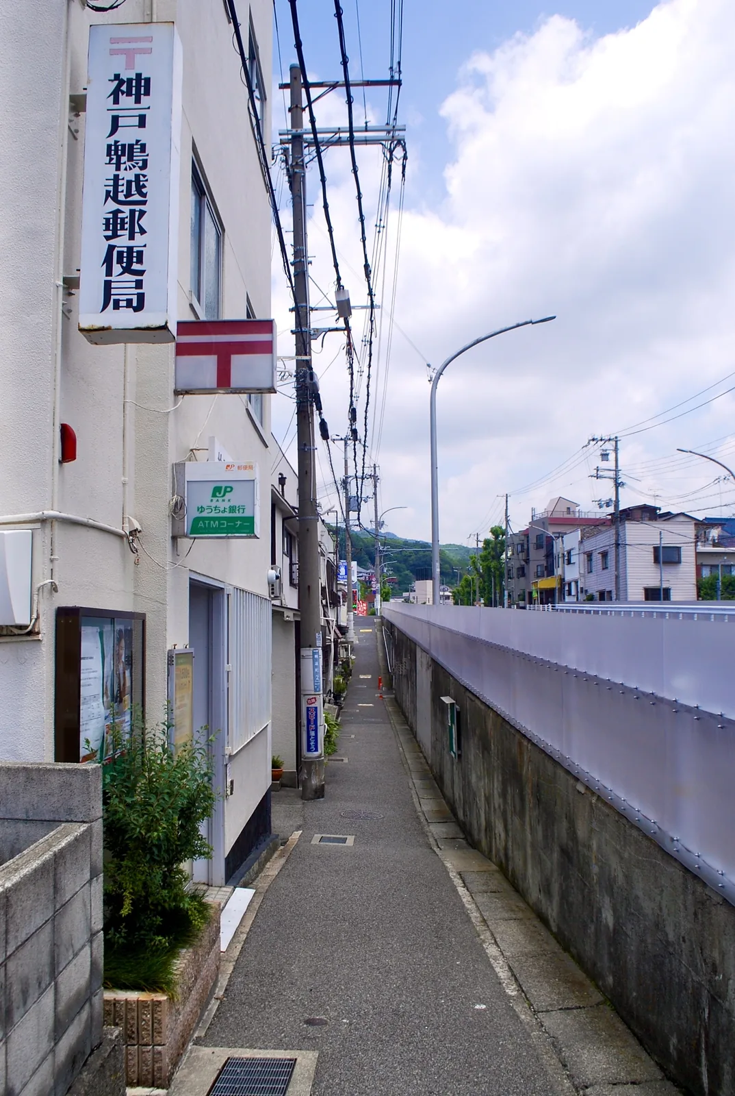 縦長の郵便局の看板と、高い壁に沿ってまっすぐ続く細い歩道