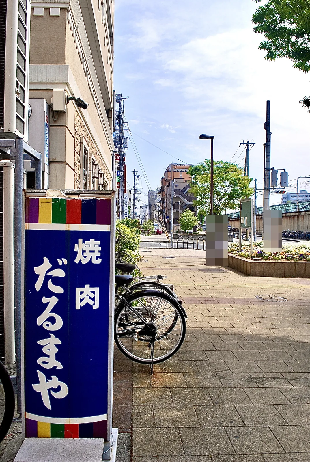 青い「焼肉 だるまや」の縦看板と、花壇のある広場