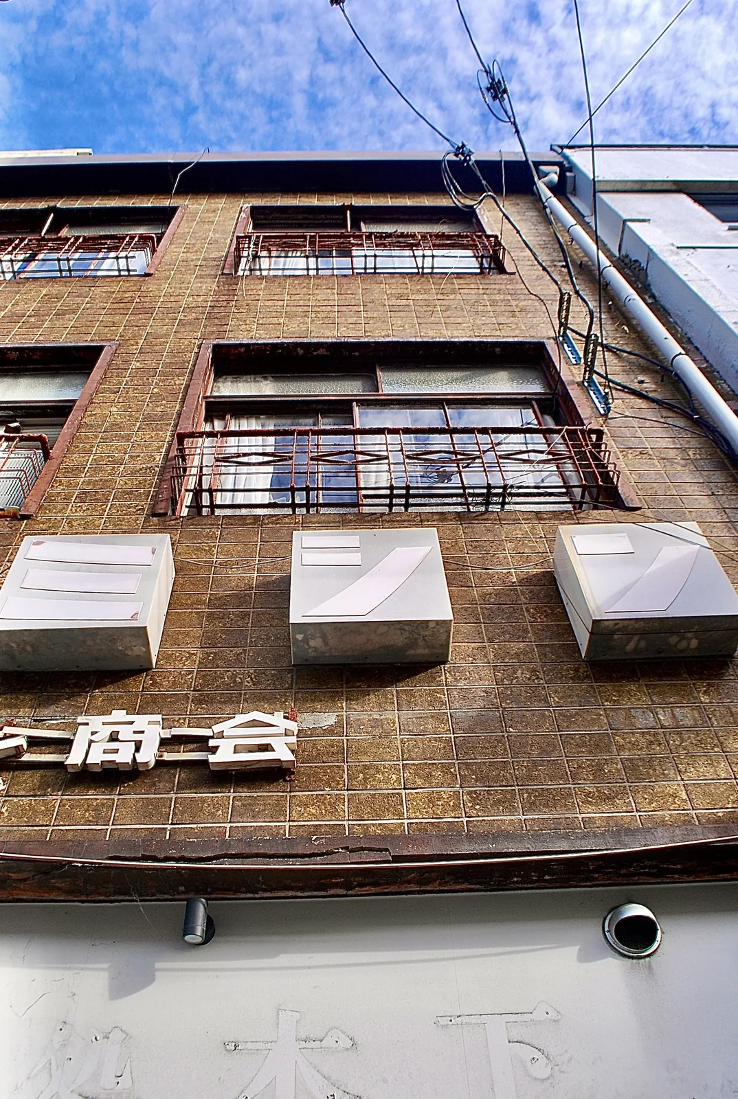 茶色いタイル張りの壁に、窓の柵と「商会」の文字が残る古いビル
