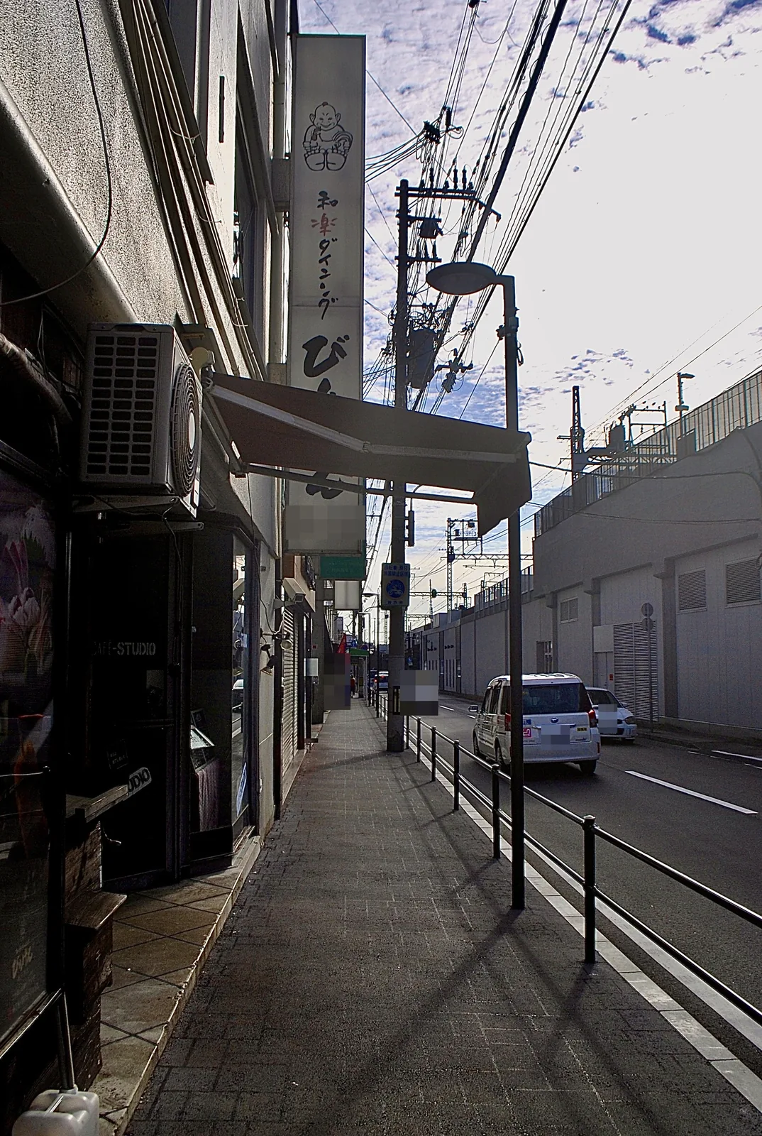 店の日よけと電柱の並ぶ歩道と、線路の高架沿いの道路