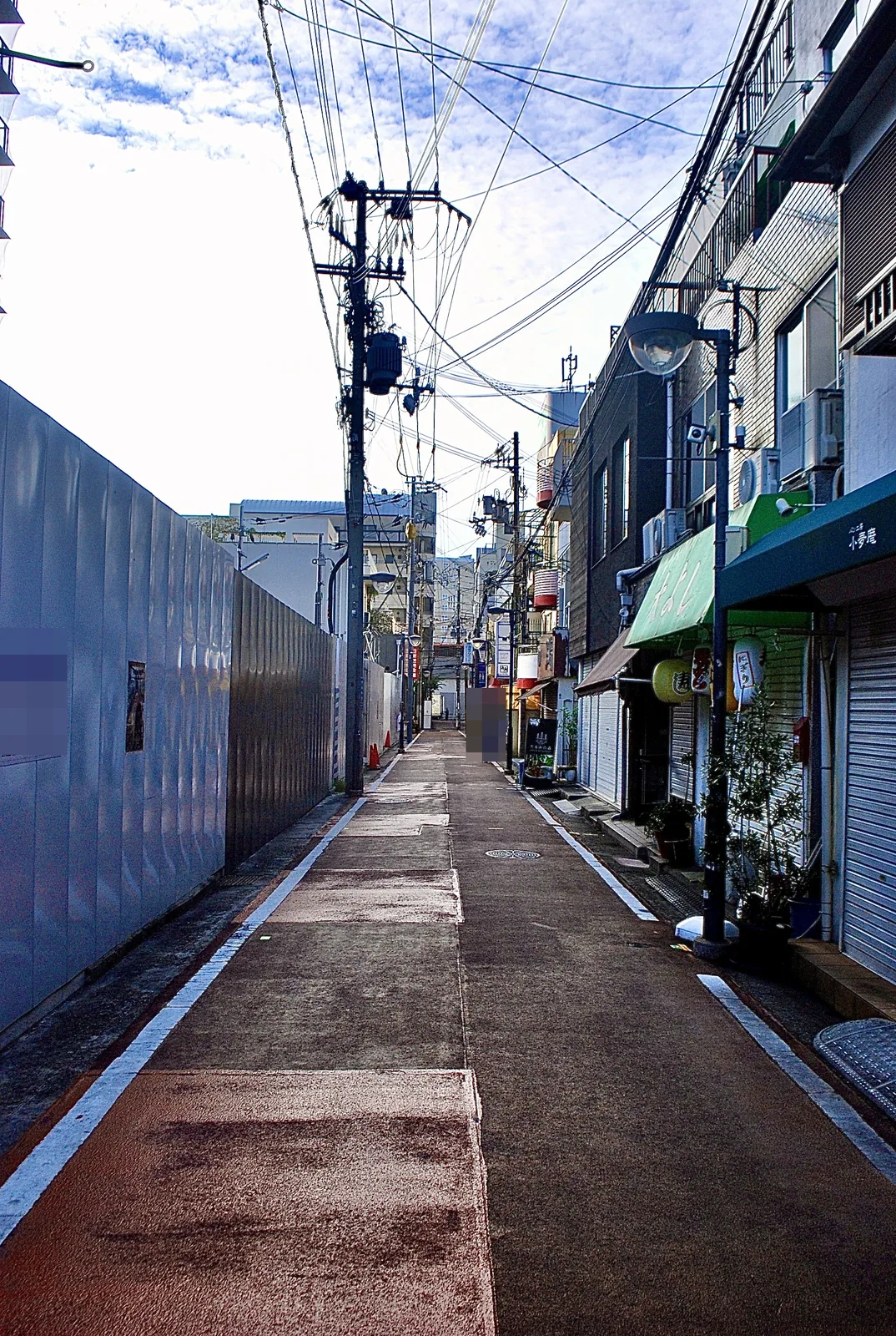 白い工事の囲いと小さな店のあいだを、まっすぐ続く赤い舗装の道