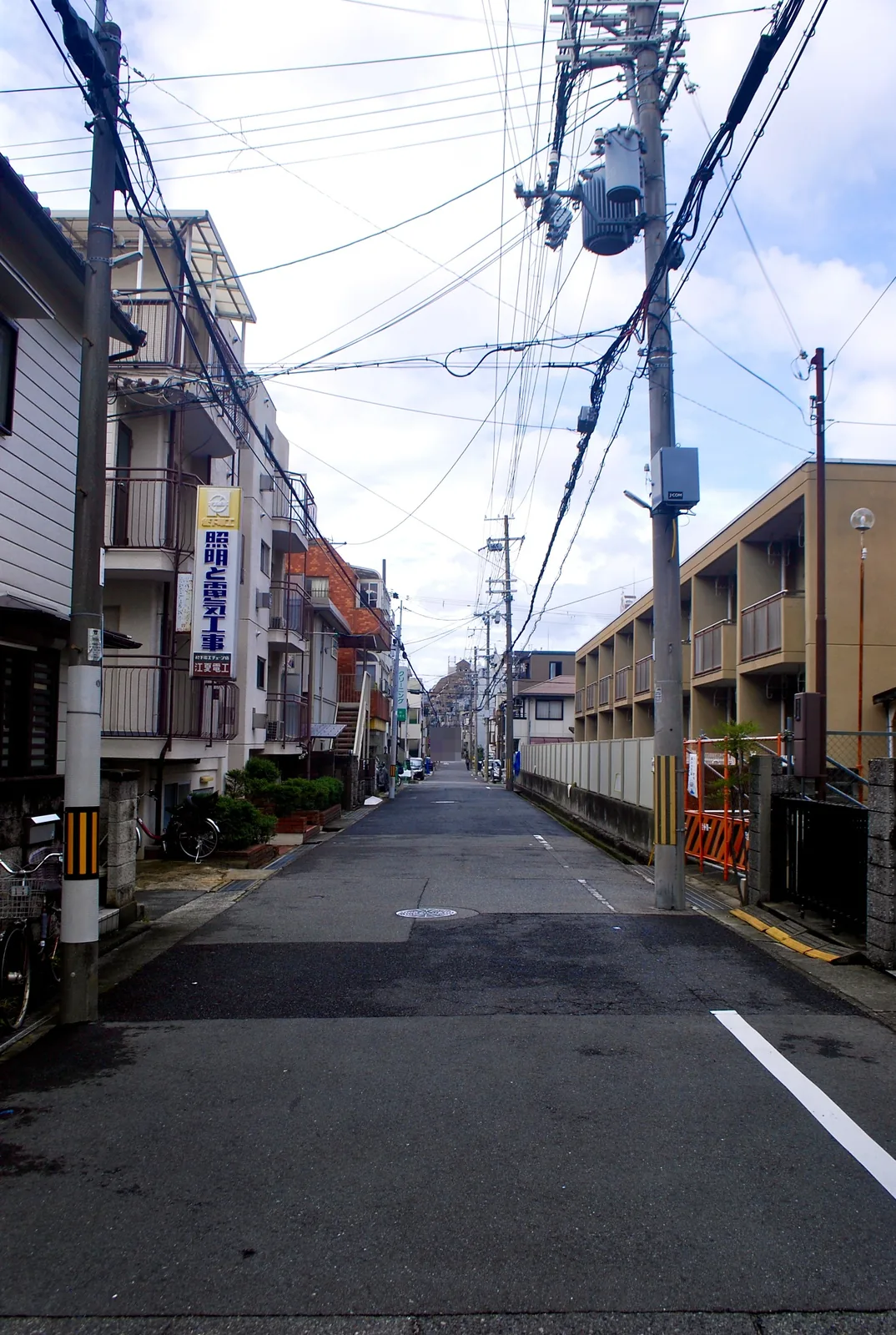 「照明と電気工事」の看板の先に、まっすぐ続く住宅街の道