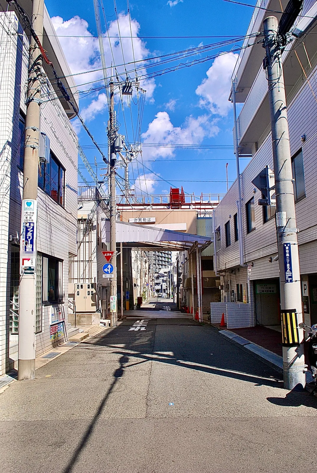 電柱の看板が並び、奥に商店街の入口が見える青空の下の通り