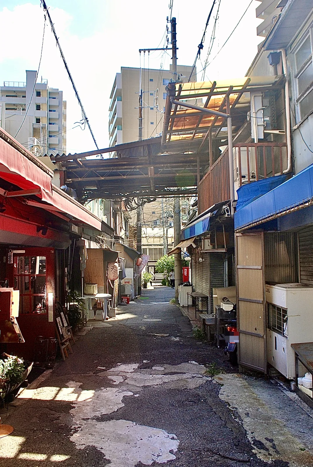 赤や青の日よけの店先が並ぶ、ひび割れた舗装の裏通り