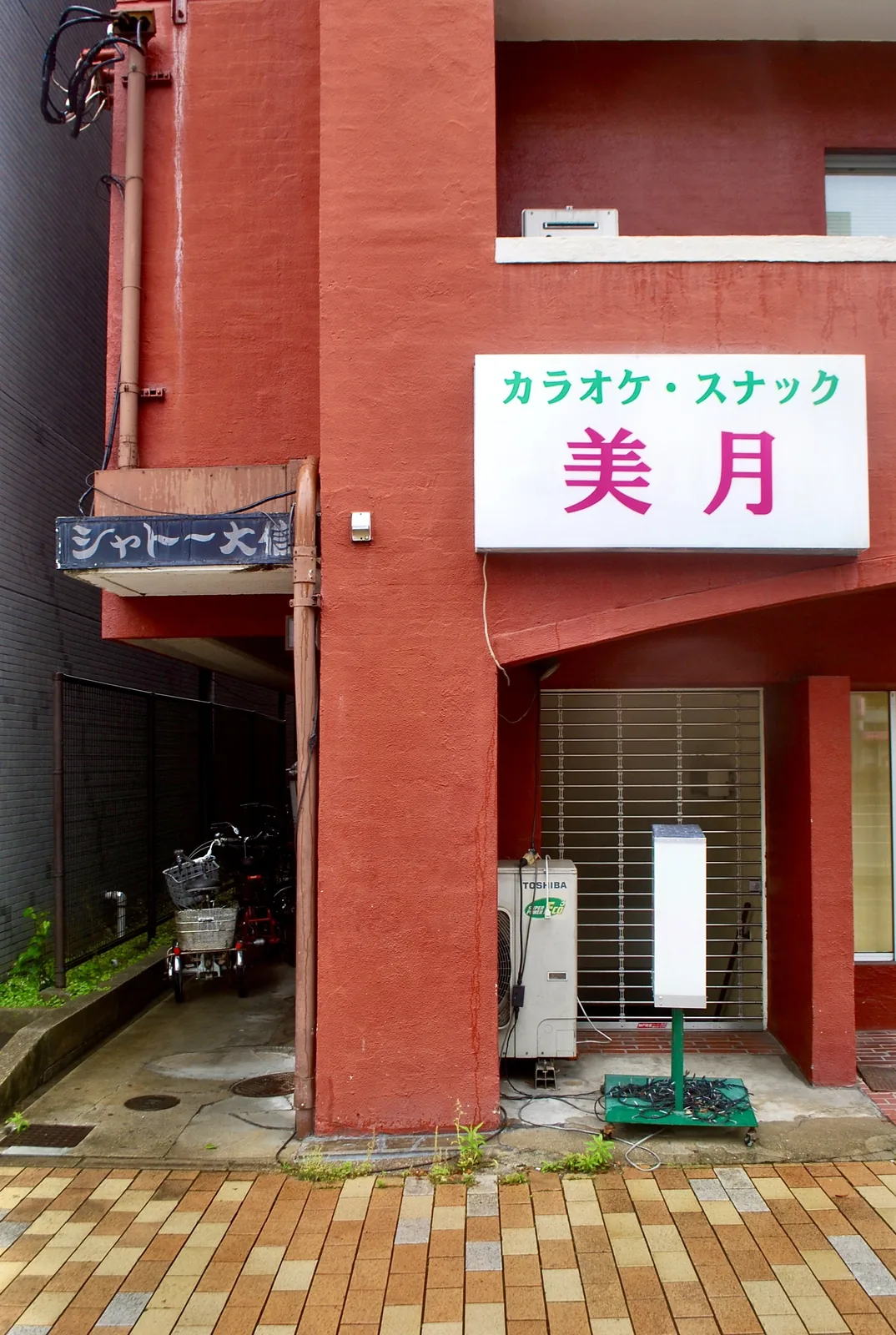 赤い壁の古いマンションの入口と、看板の残る軒