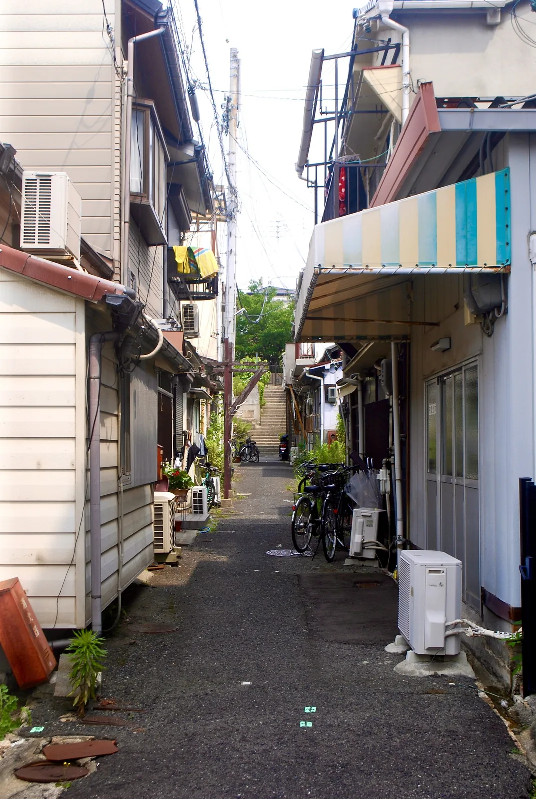 しましまの日よけと自転車の並ぶ、奥の階段へ続く細い路地