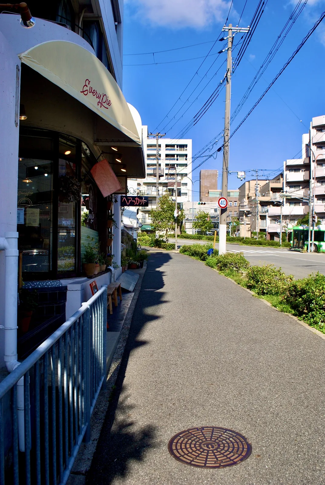 白い日よけのパン屋の店先と、植え込みの続く明るい歩道