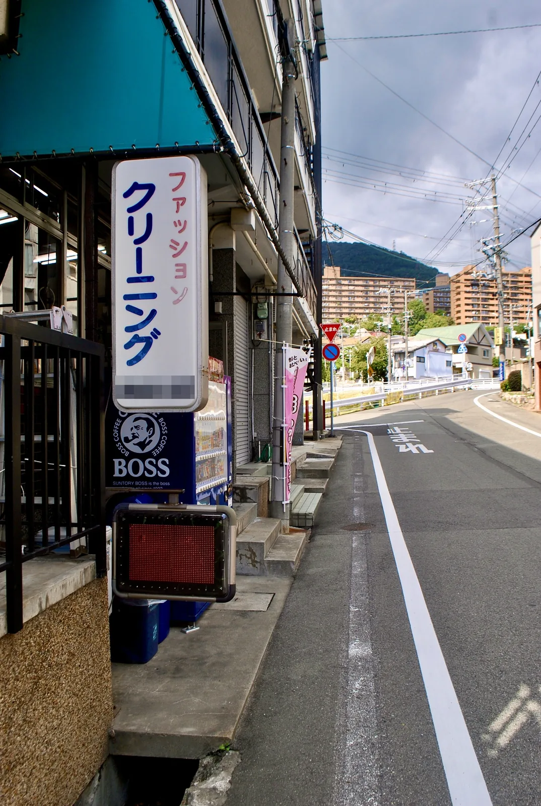 古い縦長の看板と自動販売機の並ぶ店先の先に、山と町が見える通り