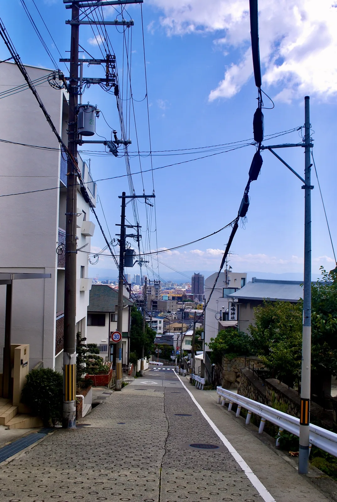 電柱と電線が並ぶ坂道の先に、遠くの町並みが見える風景