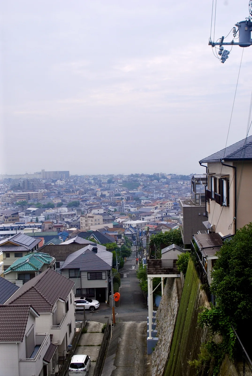 急な坂の上から、段々に重なる屋根と遠くの街並みを見る風景